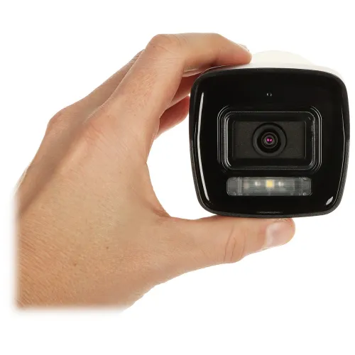 Zdjęcie przedstawia rękę trzymającą kamerę monitoringu z widocznym obiektywem i diodami LED. Kamera ma czarny front i białą obudowę.