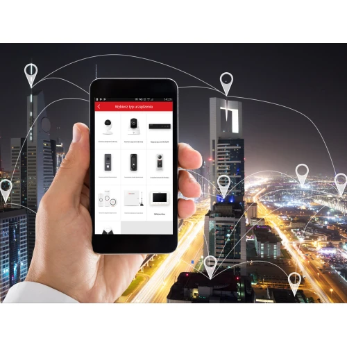 Na zdjęciu widać smartfon z aplikacją do zarządzania urządzeniami smart home, trzymany na tle nocnej panoramy miasta z zaznaczonymi lokalizacjami. Aplikacja pokazuje różne urządzenia, takie jak kamery i czujniki, co sugeruje ich integrację z systemem monitoringu.