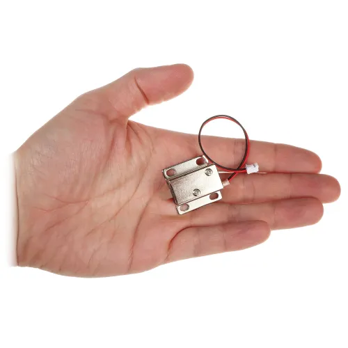 Osoba trzyma w dłoni mały, metalowy zamek elektromagnetyczny z dwoma przewodami zakończonymi białą wtyczką. Zamek ma prostokątny kształt i cztery otwory montażowe.