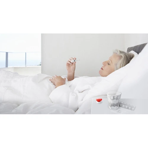 Na zdjęciu znajduje się niebieski przycisk Fibaro, umieszczony na białym stoliku obok szklanki wody i blistra z tabletkami. Produkt jest kompaktowy i ma minimalistyczny design, idealny do nowoczesnych wnętrz.