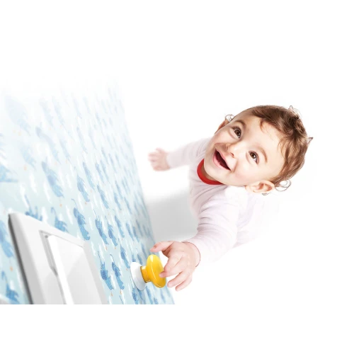 Na zdjęciu znajduje się niebieski przycisk Fibaro, zamontowany na ścianie z wzorem w niebieskie wieloryby. Przycisk jest używany przez dziecko, które sięga do niego z uśmiechem.