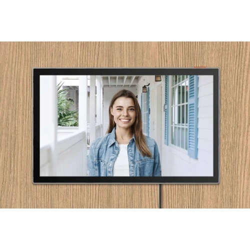 Na zdjęciu widać ekran elektronicznego wizjera drzwiowego, na którym wyświetlana jest kobieta stojąca na zewnątrz domu. Ekran jest umieszczony na drewnianej powierzchni.