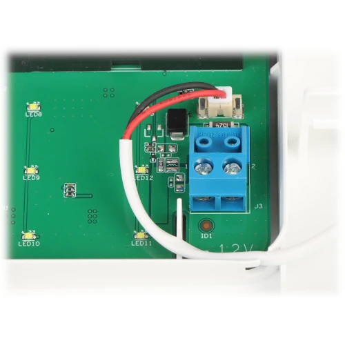 Zdjęcie przedstawia zieloną płytkę drukowaną z niebieskim złączem śrubowym, do którego podłączone są czerwony i czarny przewód. W tle widoczne są różne elementy elektroniczne, takie jak diody LED i rezystory.